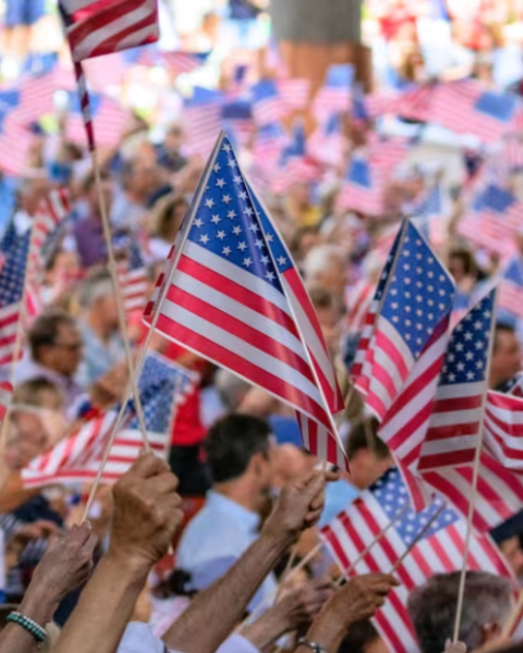 A crowd of people waving American flags, creating a festive and patriotic atmosphere at what seems like a public gathering or event.