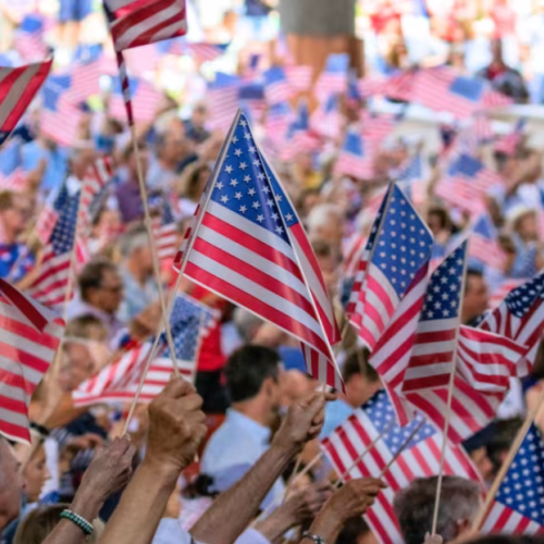 A crowd of people waving American flags, creating a festive and patriotic atmosphere at what seems like a public gathering or event.