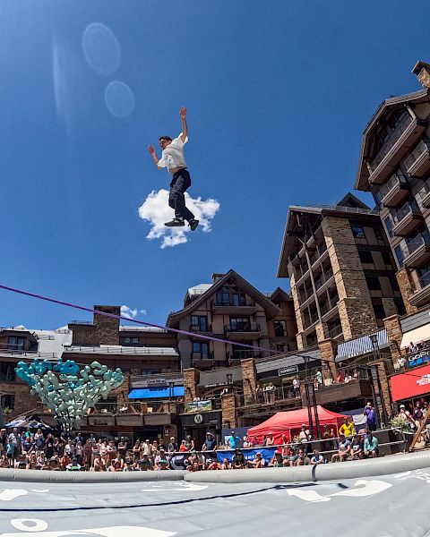 A person is performing a high jump onto an inflatable airbag in a busy outdoor setting with buildings and a crowd below.