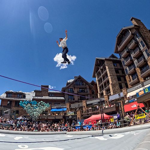 A person is performing a high jump onto an inflatable airbag in a busy outdoor setting with buildings and a crowd below.