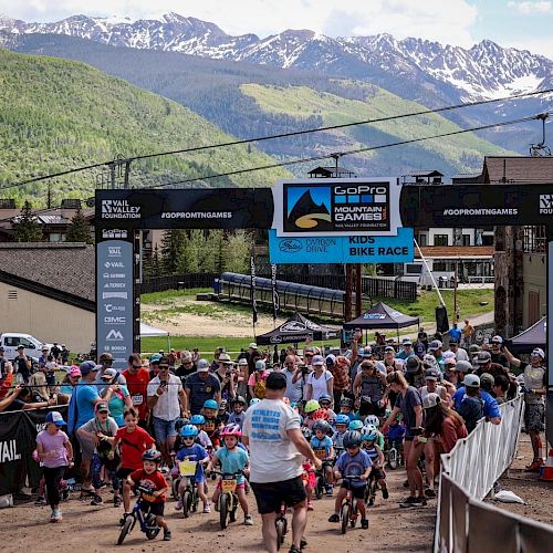 A group of children and adults gather at a starting line for a bike race event in a mountainous area with banners and a lively atmosphere.