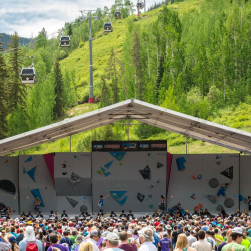 An outdoor climbing competition with spectators, a climbing wall, cable cars, and forested hills in the background under a blue sky.