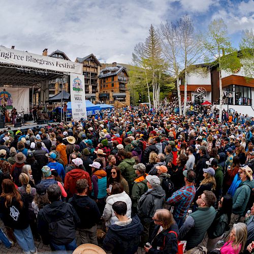 A large crowd gathers at an outdoor festival with a stage performing, buildings in the background, and a bright, partly cloudy sky.