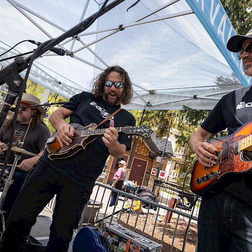 A live outdoor band performs under a canopy; two guitarists strum electric/acoustic guitars while others play nearby, crowds watching.