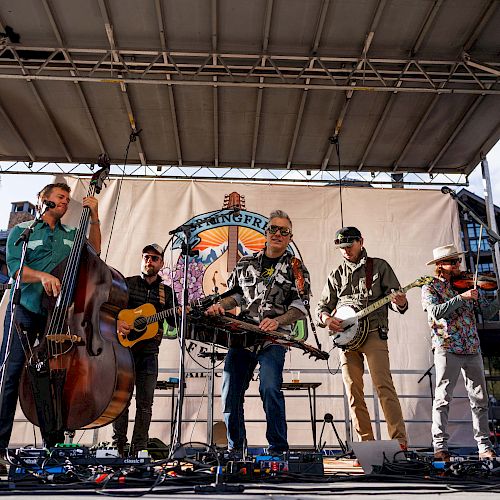 A group of musicians performing on an outdoor stage with various instruments, and a banner backdrop displaying colorful graphics and text.