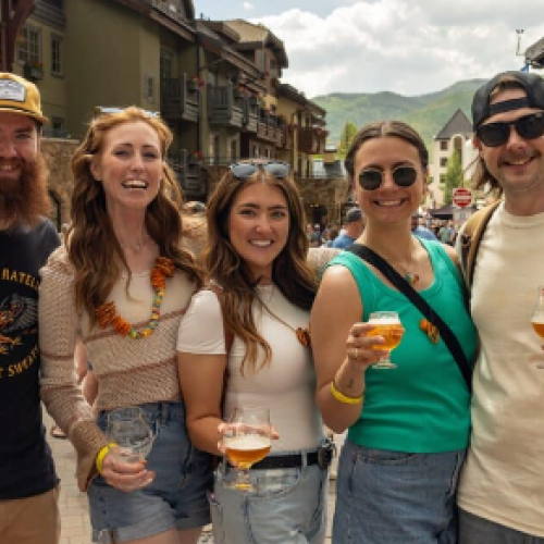 A group of five friends posing outdoors, smiling with beer glasses, mountains in the background, sunny day, casual summer outfits.