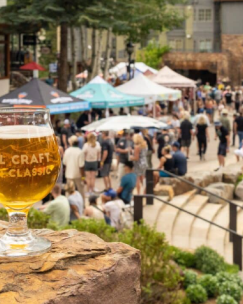 A glass of beer sits on a rock with a crowd and tents in the background at an outdoor event.