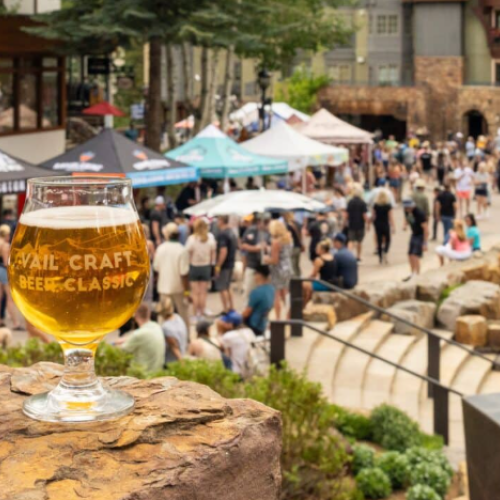 A glass of beer sits on a rock with a crowd and tents in the background at an outdoor event.