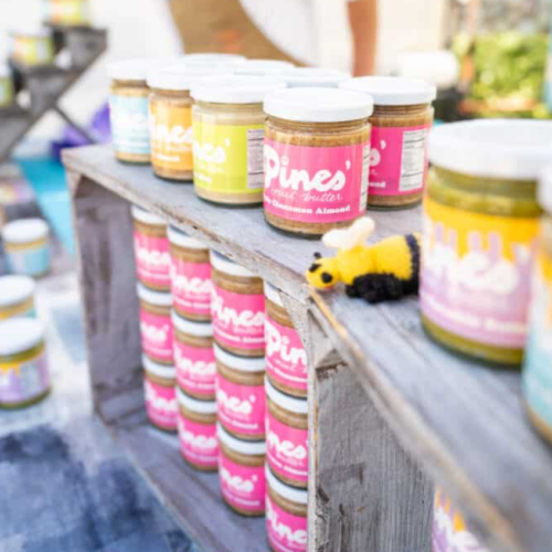 Jars of nut butter are displayed on wooden shelves, alongside a small toy bee decoration, creating a vibrant market stall scene.