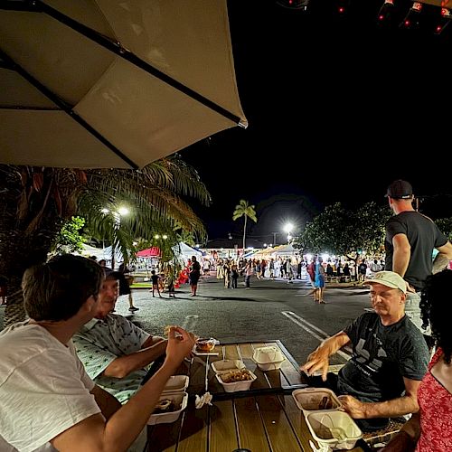 People dining at a night market or seaside food stall, with friends chatting under a large umbrella as others wander the lit street.