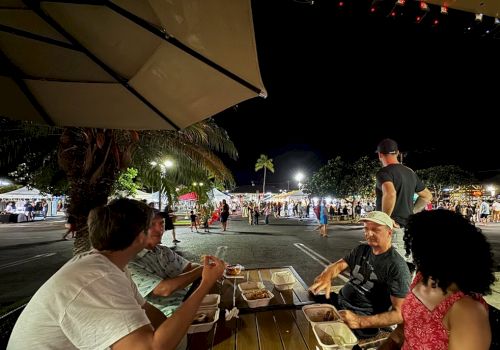 People are dining under an umbrella at night, with a lively outdoor market scene in the background.