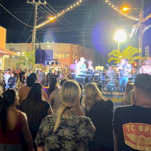 A crowd watches a band performing outdoors at night, with string lights overhead and palm trees visible in the background.