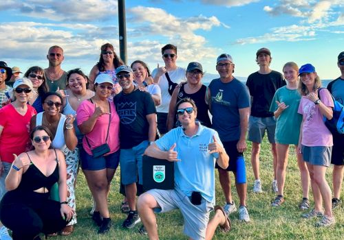 A group of people posing outdoors on grass under a blue sky, some with thumbs up and wearing casual attire, with a scenic background.