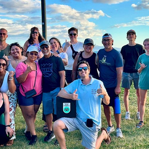 A group of people posing outdoors on grass under a blue sky, some with thumbs up and wearing casual attire, with a scenic background.