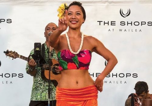 A woman performs a hula dance with musicians in the background at The Shops at Wailea, wearing a floral outfit and lei.