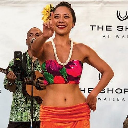 A woman performs a hula dance with musicians in the background at The Shops at Wailea, wearing a floral outfit and lei.