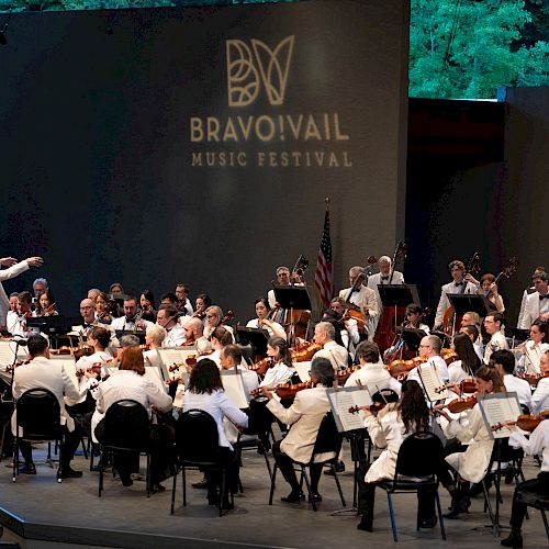 An orchestra performs on stage at the Bravo! Vail Music Festival, with a conductor leading musicians dressed in white.