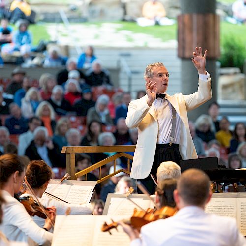 A conductor leads an orchestra with musicians, performing outdoors before an audience seated in the background.