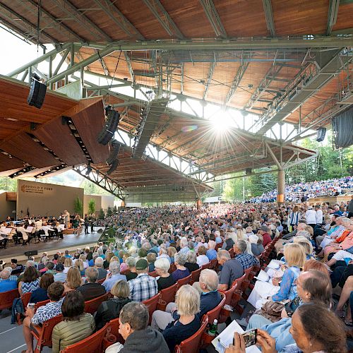 A large outdoor amphitheater with a full audience, a stage with an orchestra, and sunlight streaming through the roof structure.