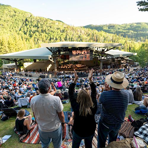 The image shows an outdoor concert in a scenic, forested area with a large audience seated on the grass, enjoying the performance.