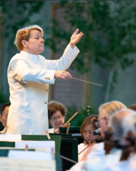 A conductor in a white coat leads an outdoor orchestra performance, with musicians focused on playing their instruments.