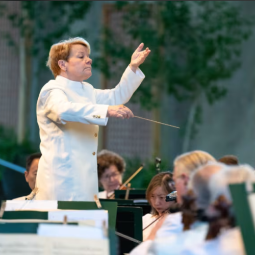 A conductor in a white coat leads an outdoor orchestra performance, with musicians focused on playing their instruments.