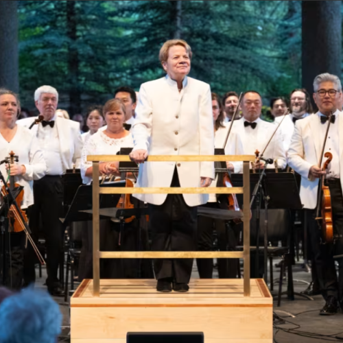 A conductor in a white jacket stands before a large choir/orchestra on stage, surrounded by musicians in white shirts and instruments.