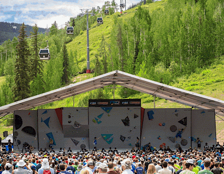 A large outdoor rock climbing competition is underway under a shaded tent stage, with a big crowd watching as gondola lifts ascend a green hillside.