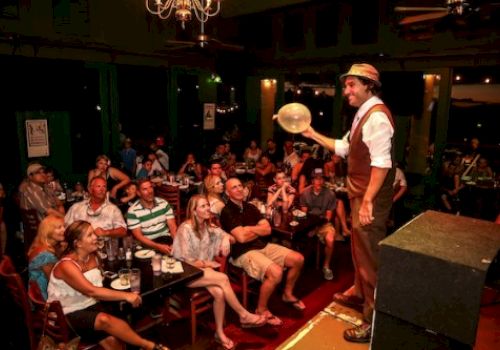 A performer on stage entertains a seated audience with a balloon trick at a lively nighttime indoor venue.
