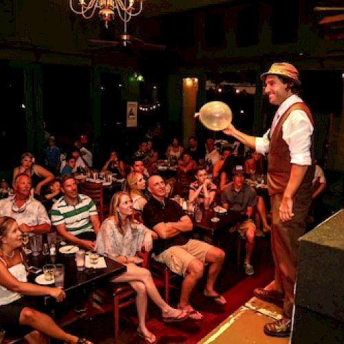 A performer on stage entertains a seated audience with a balloon trick at a lively nighttime indoor venue.