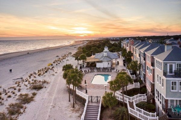Beachfront view at sunset with a pool, houses, and a pavilion on the shore; vibrant skies and sandy beach stretch into the distance.