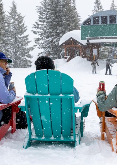 Three people in colorful chairs in the snow, next to a snowboard, watching a ski lift.