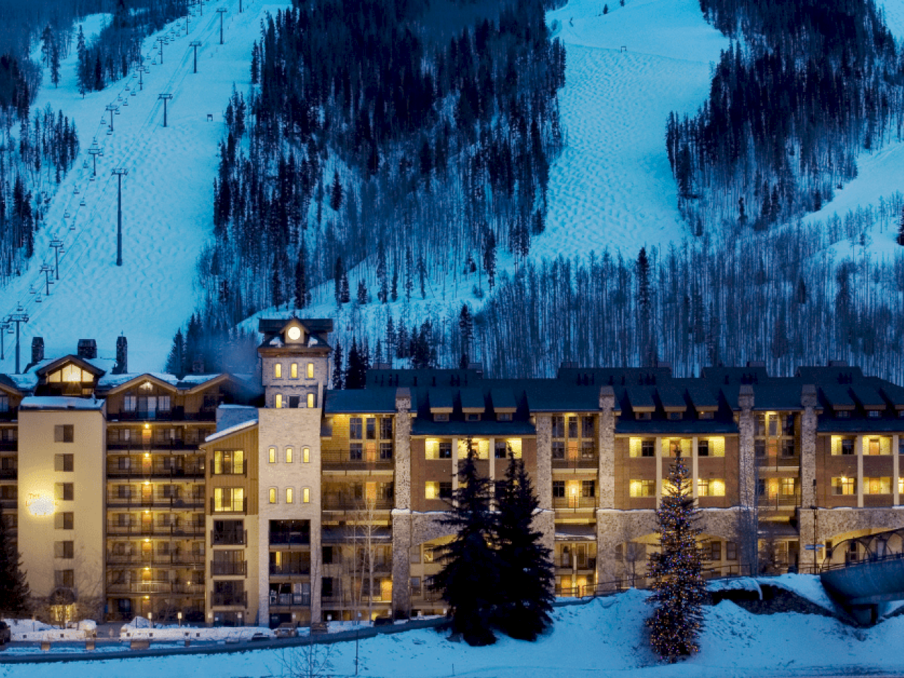A large hotel lit up at night in a snowy landscape, with ski slopes and trees in the background, creating a serene winter scene.
