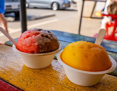 The image shows two colorful shaved ice treats in cups with spoons, placed on a colorful outdoor table with cars and people in the background.