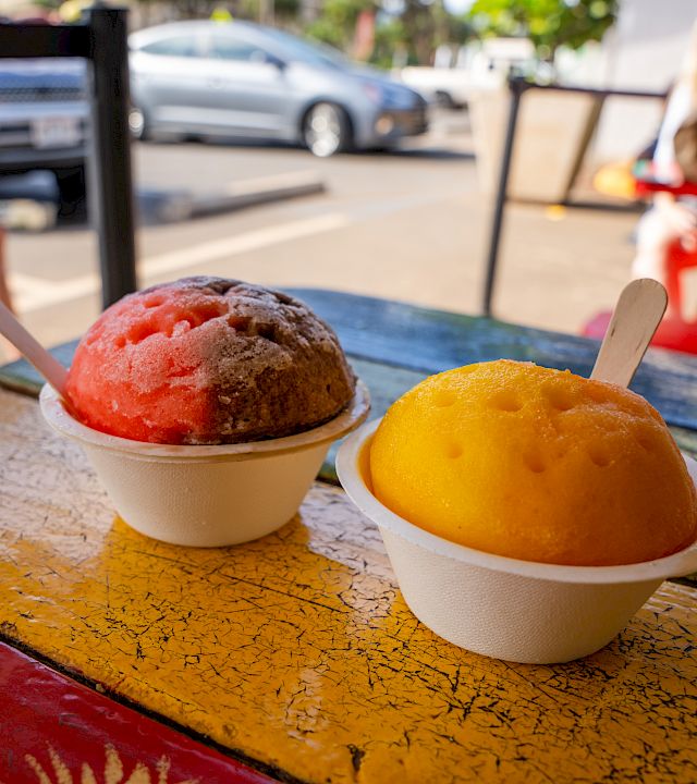The image shows two colorful shaved ice treats in cups with spoons, placed on a colorful outdoor table with cars and people in the background.