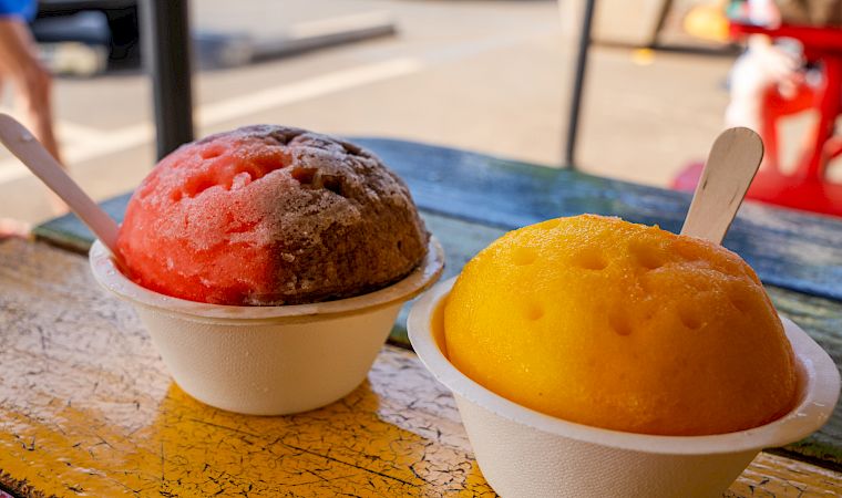 The image shows two colorful shaved ice treats in cups with spoons, placed on a colorful outdoor table with cars and people in the background.