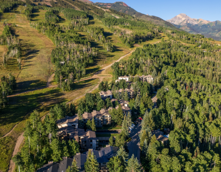 Aerial view of a mountainous landscape with a forested area, resorts, and winding trails, under a clear blue sky and distant peaks.