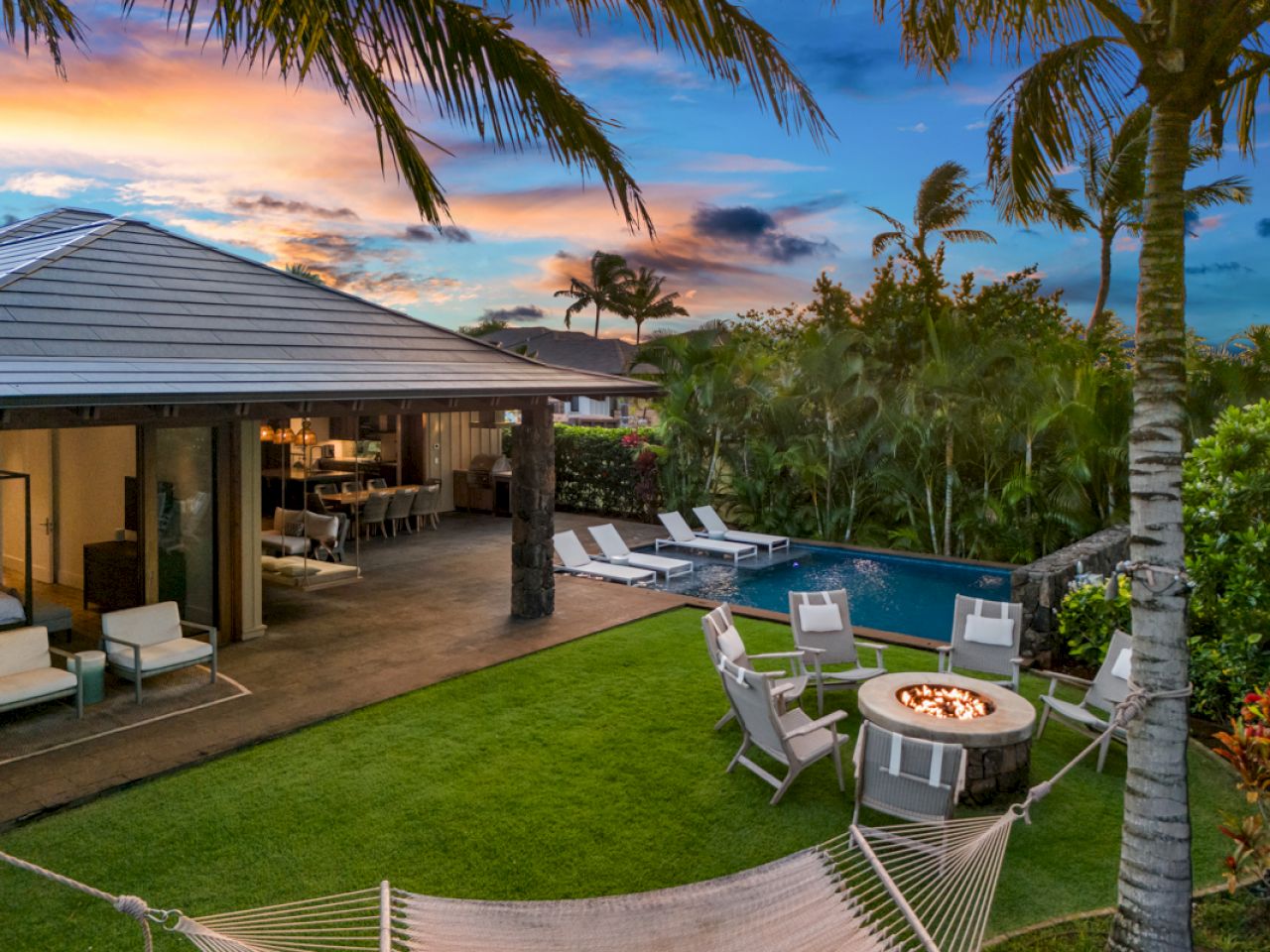 A tropical backyard features a pool, fire pit with chairs, hammock, and outdoor seating under a sunset sky.