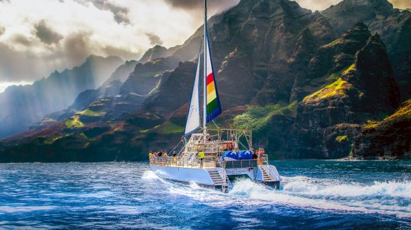 A sailboat is navigating vibrant blue waters near towering, rugged mountains under a sky with scattered clouds and rays of sunlight.