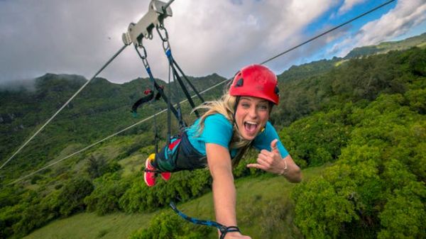 A person is zip-lining over a lush landscape, wearing a red helmet and blue shirt, displaying a joyful expression and hand gesture.