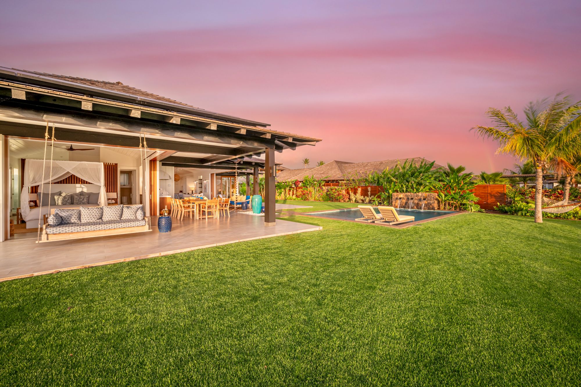 The image shows a luxurious outdoor patio with a swing, dining area, and pool, set against a vibrant pink sunset sky and landscaped lawn.