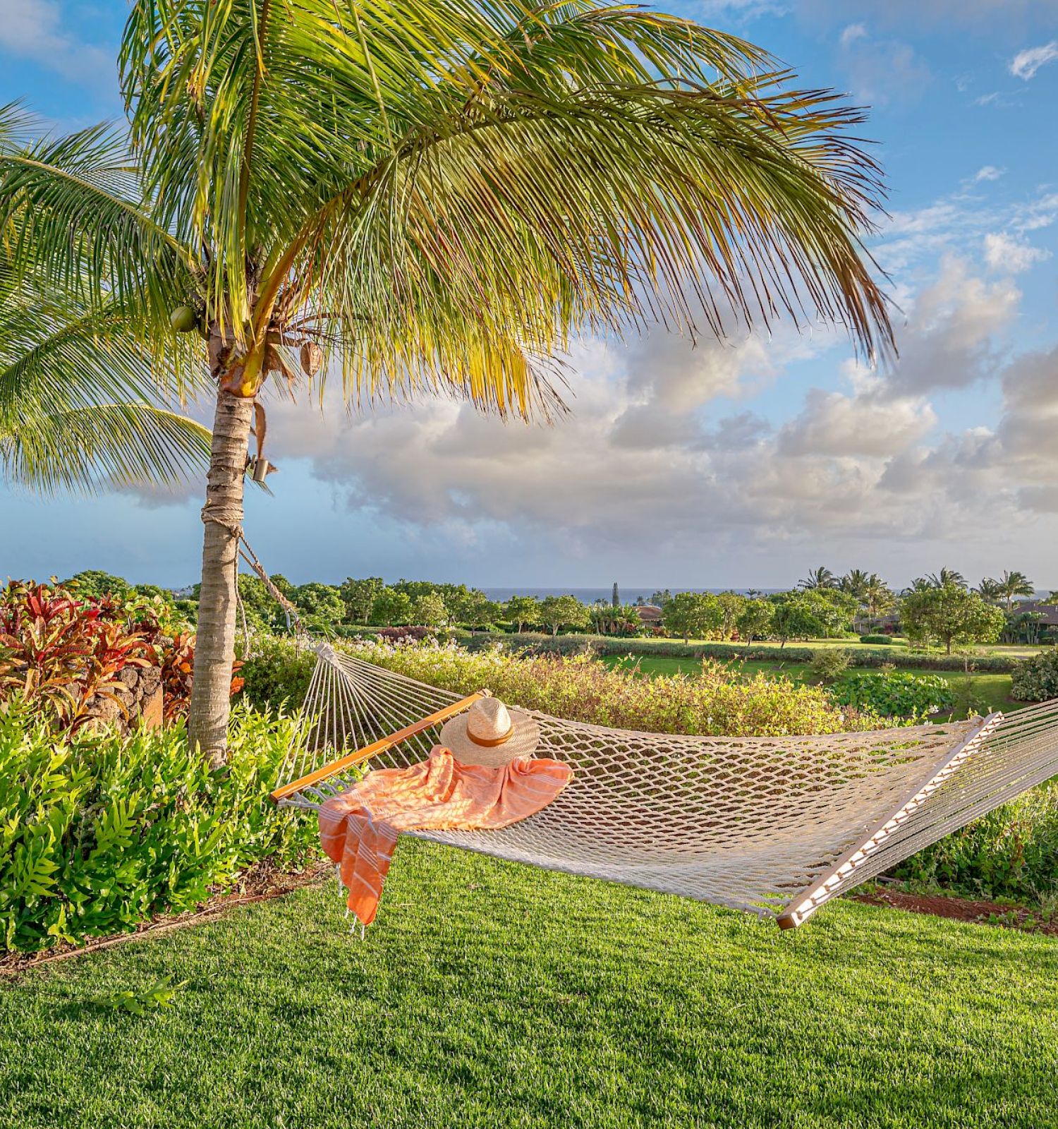 A hammock with a person wearing a hat and dress is set between palm trees in a lush garden with a scenic view of the sky and clouds.