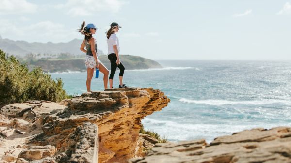 Two people stand on a rocky cliff overlooking the ocean, with a scenic view and clear skies in the background.
