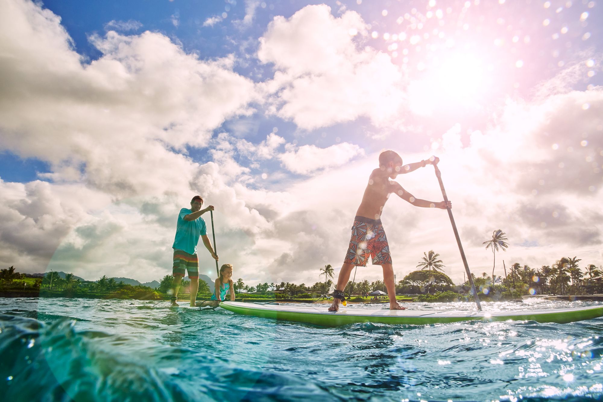 People paddleboarding on a sunny day with palm trees in the background, under a partly cloudy sky.