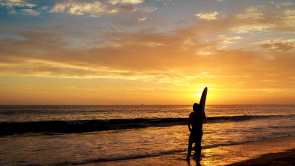 A person holding a surfboard stands on a beach during a vibrant sunset, with waves gently lapping at the shore.