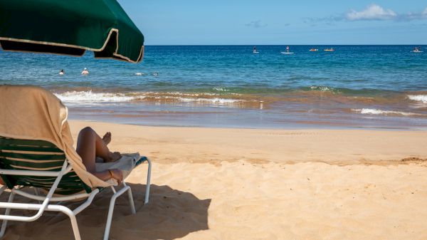 A person relaxes on a beach chair under an umbrella, facing the ocean waves, with kayakers visible in the distance on a sunny day.