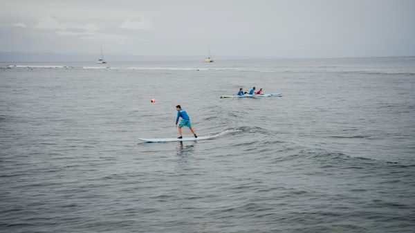 A person is stand-up paddleboarding on the sea, with a distant canoe and buoys nearby, under a cloudy sky.