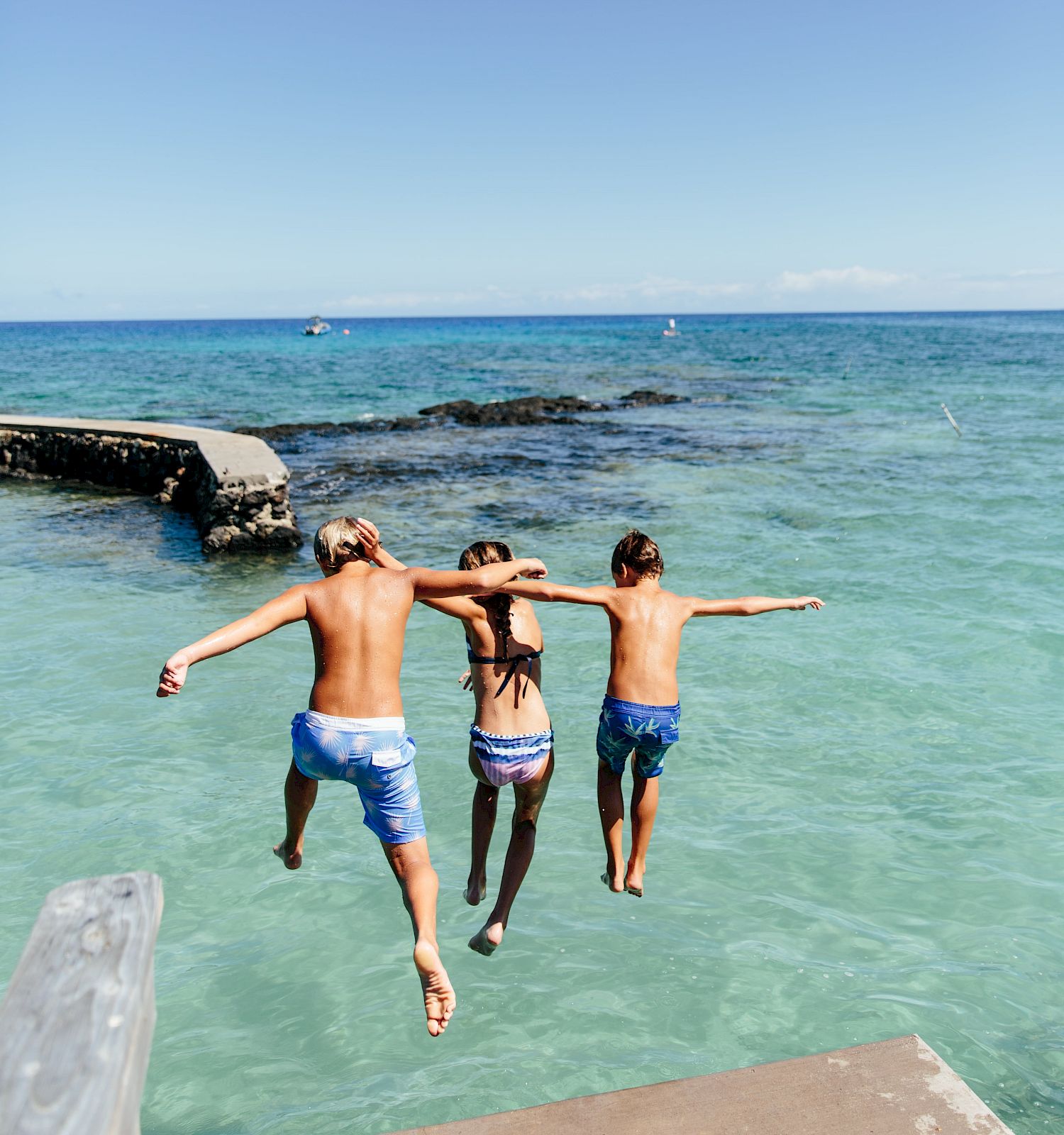 Three people are jumping into clear blue ocean water from a ledge, under a bright blue sky with scattered clouds.