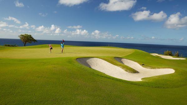 A scenic golf course by the ocean, with two people on the green, a sand trap in the foreground, under a blue sky with scattered clouds.