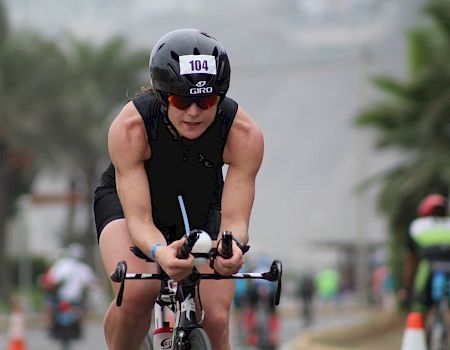A cyclist wearing a helmet and bib number is riding on a road during a race, with blurred participants and palm trees in the background.
